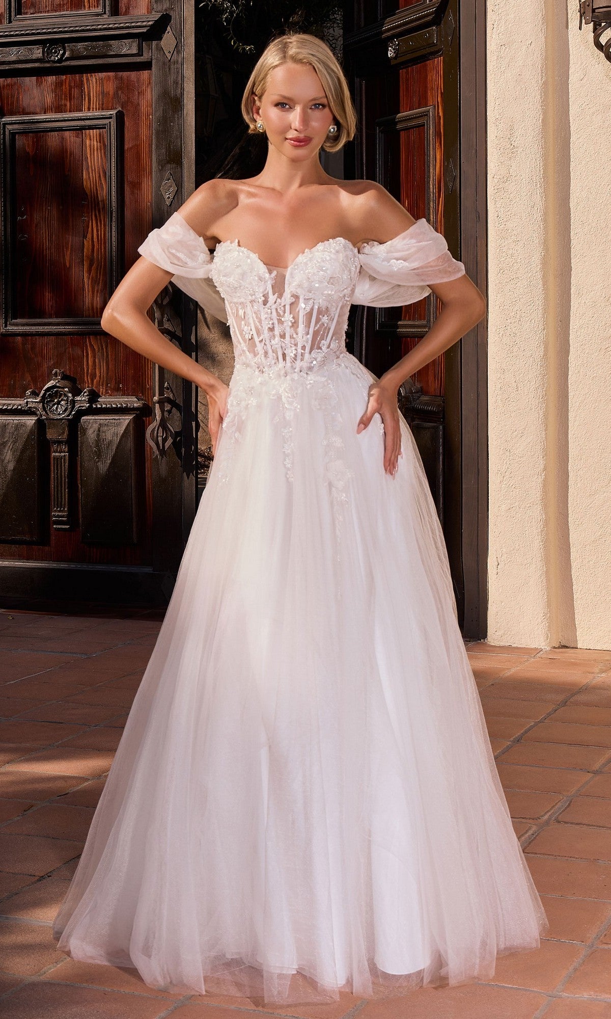 A woman in the Ladivine CD2426W long formal dress, featuring an off-the-shoulder, off-white tulle design with a fitted illusion corset bodice and floral details, stands before wooden doors.